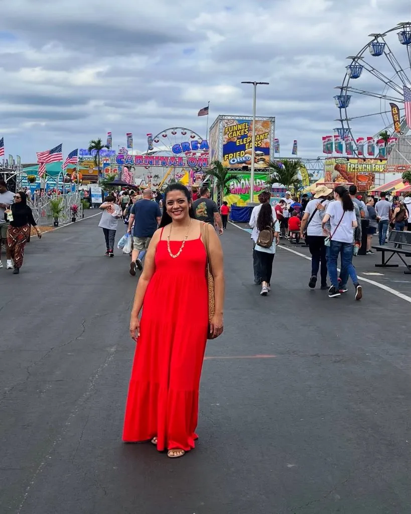 Woman wearing a red maxi dress and sandals at a summer county fair surrounded by carnival rides and food stands. Cute fair outfit summer idea for moms.