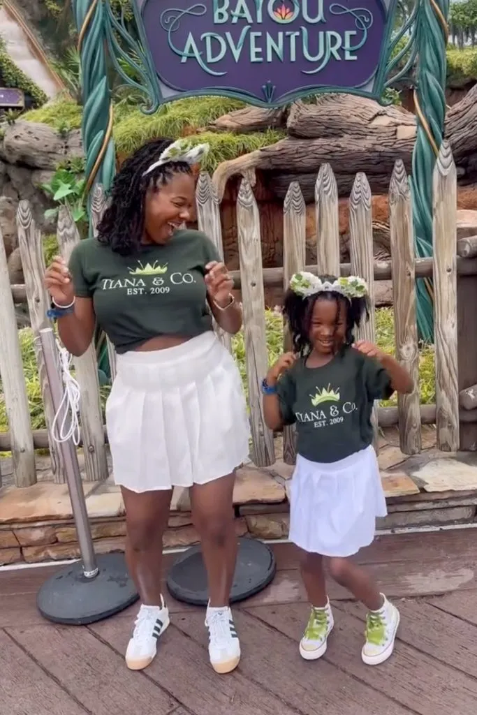 Mom and daughter wearing matching green “Tiana & Co.” shirts with white skirts and sneakers in front of Tiana’s Bayou Adventure sign at Disney World. Example of a mommy and me summer theme park outfit for moms. Text on image says “Bayou Adventure.”