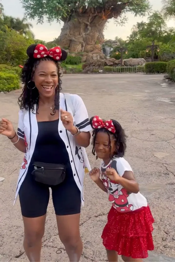 Mom and daughter in matching Minnie Mouse-inspired theme park outfits — mom in black biker shorts, Mickey jersey, and fanny pack; daughter in Minnie tee and red sparkly skirt.