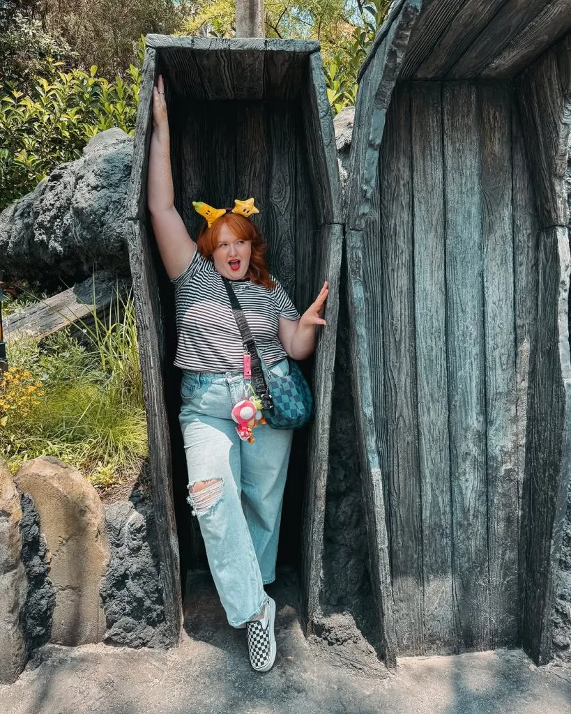 Trendy plus size amusement park outfit for moms with striped tee, distressed jeans, and checkered Vans, styled with Pokemon ears and a character crossbody.