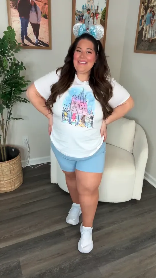 Plus size mom wearing a black skort dress and sparkly Minnie ears with white sneakers, standing indoors with Disney wall art in the background.