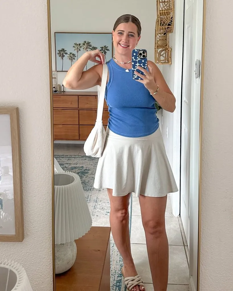 Woman taking a mirror selfie in a casual theme park outfit for moms with a fitted blue tank top, white athletic skirt, white shoulder bag, and slip-on sandals.