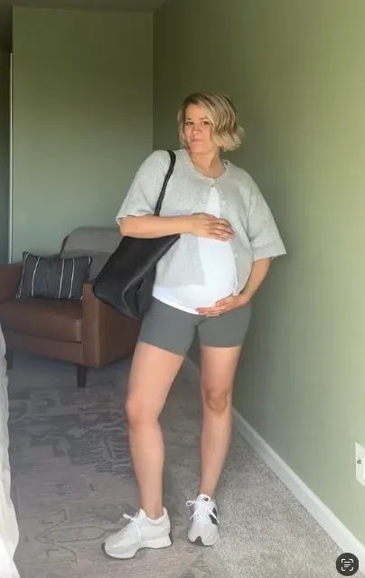 Maternity summer outfit for moms at Disney featuring a white maternity tank, open short-sleeve sweater, biker shorts, sneakers, and tote bag. Ideal maternity amusement park outfit. No text on image.