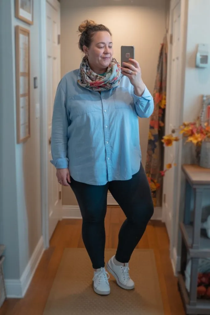 plus size woman wearing chambray shirt, black leggings, floral scarf, and white sneakers taking a mirror selfie in hallway