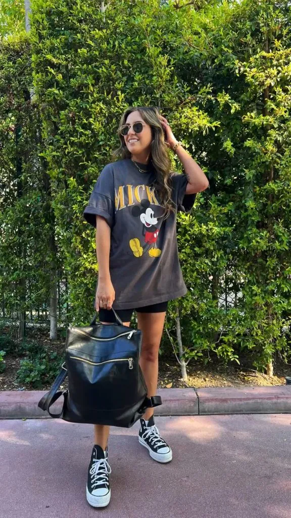 Mom wearing oversized black Mickey Mouse tee, biker shorts, Converse sneakers, and black leather backpack—edgy casual summer theme park outfit.