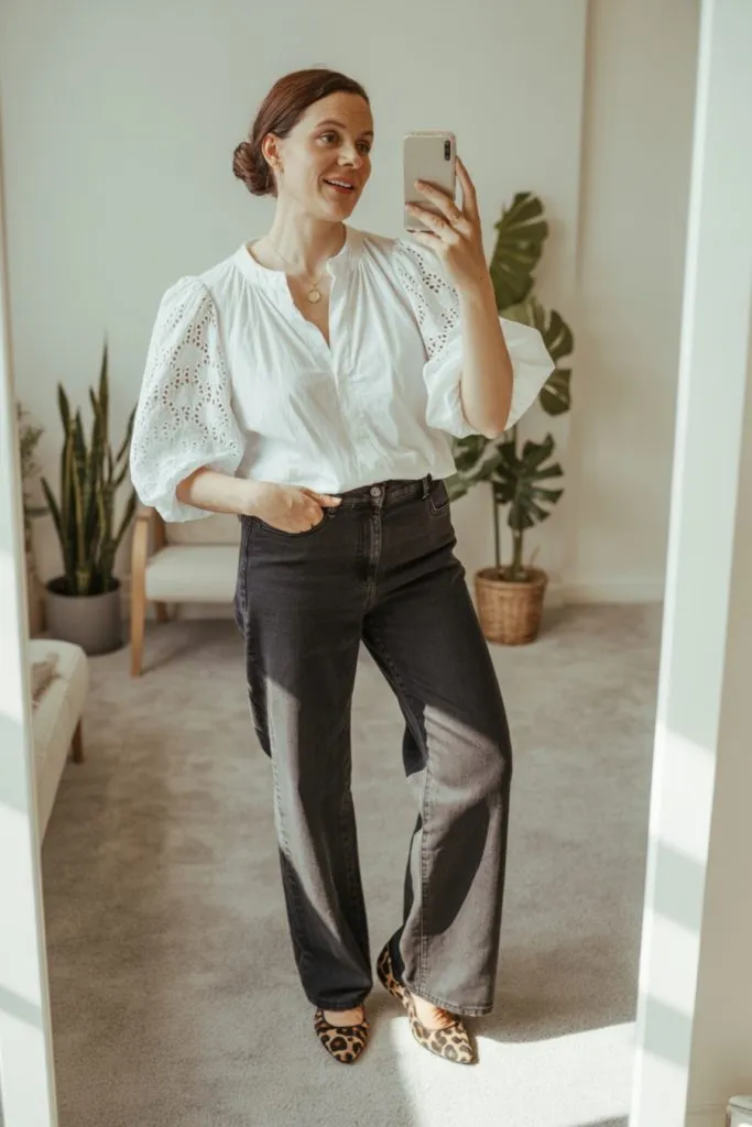 woman taking a mirror selfie in white puff-sleeve blouse, dark wash straight-leg jeans, and leopard flats