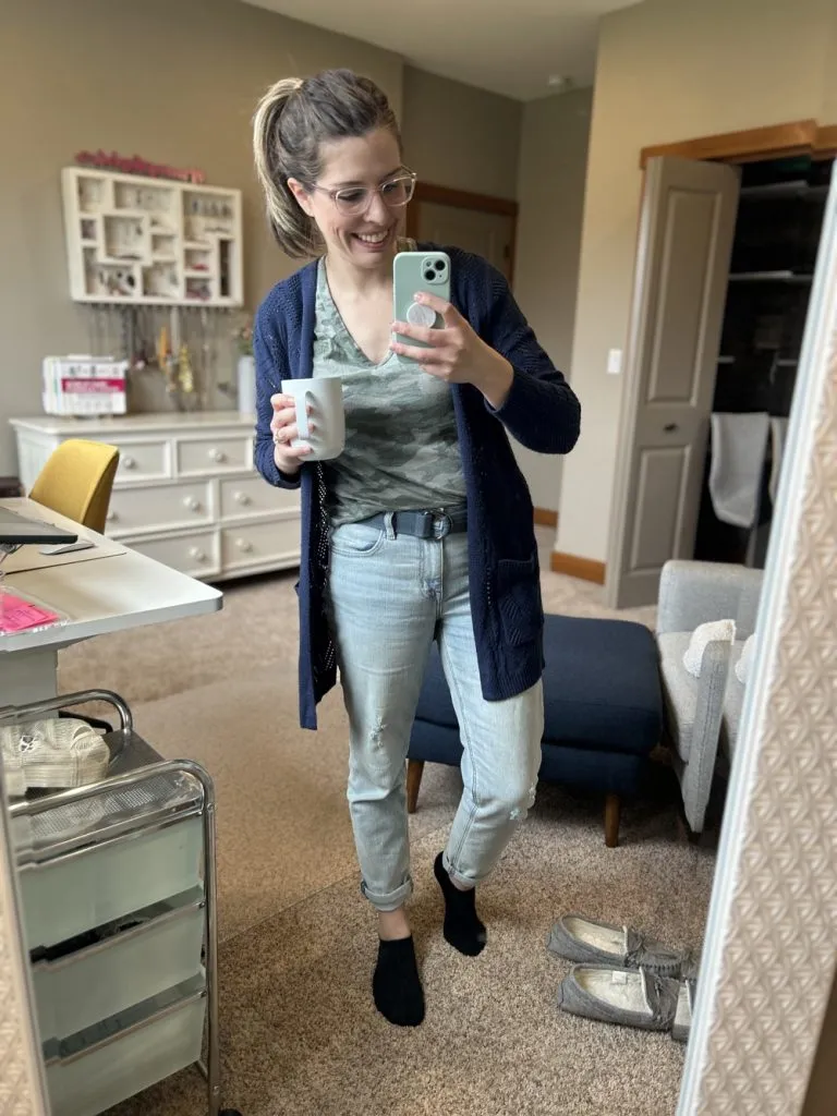 Woman wearing camo print tee, navy cardigan, and straight leg jeans, holding a coffee.