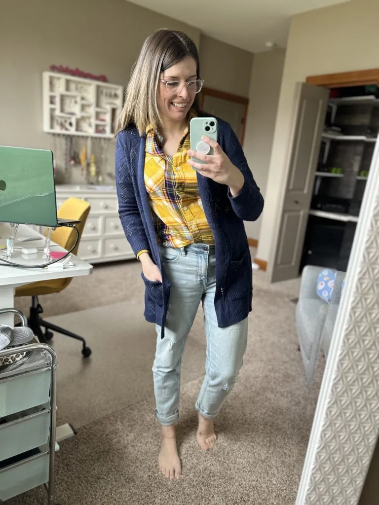 Woman wearing plaid shirt with navy cardigan and straight leg jeans, smiling in a bedroom mirror.