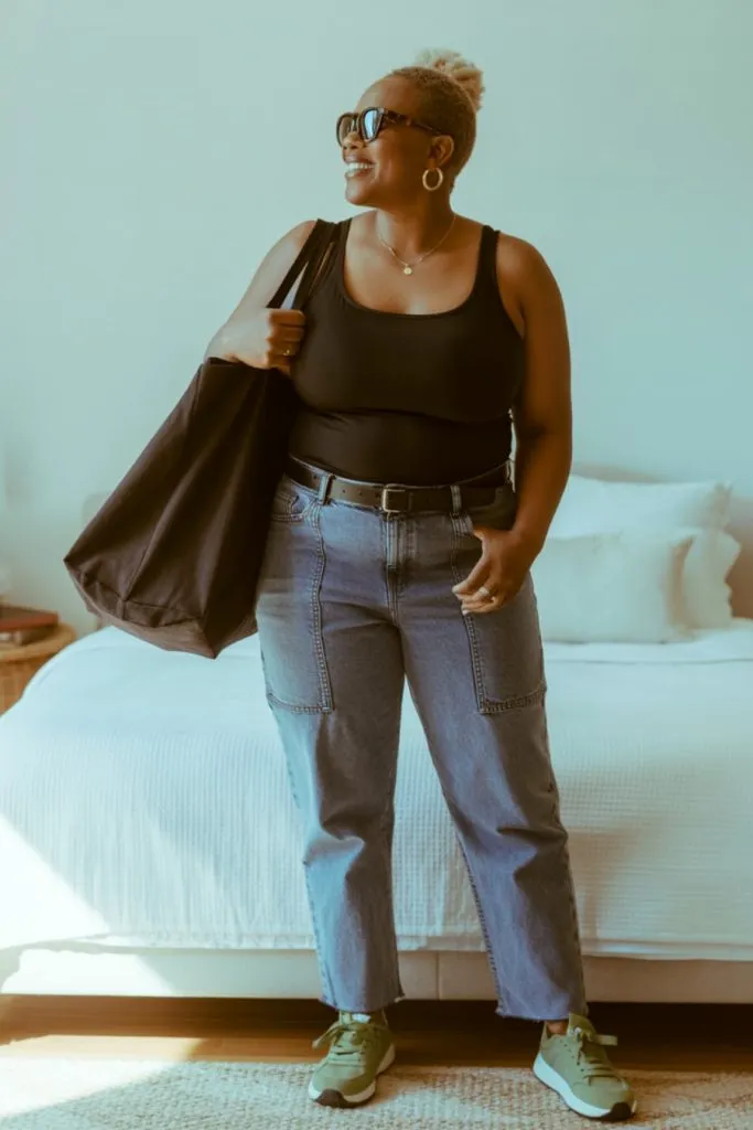 Woman in utility jeans, black tank top, and green sneakers smiling indoors with tote bag