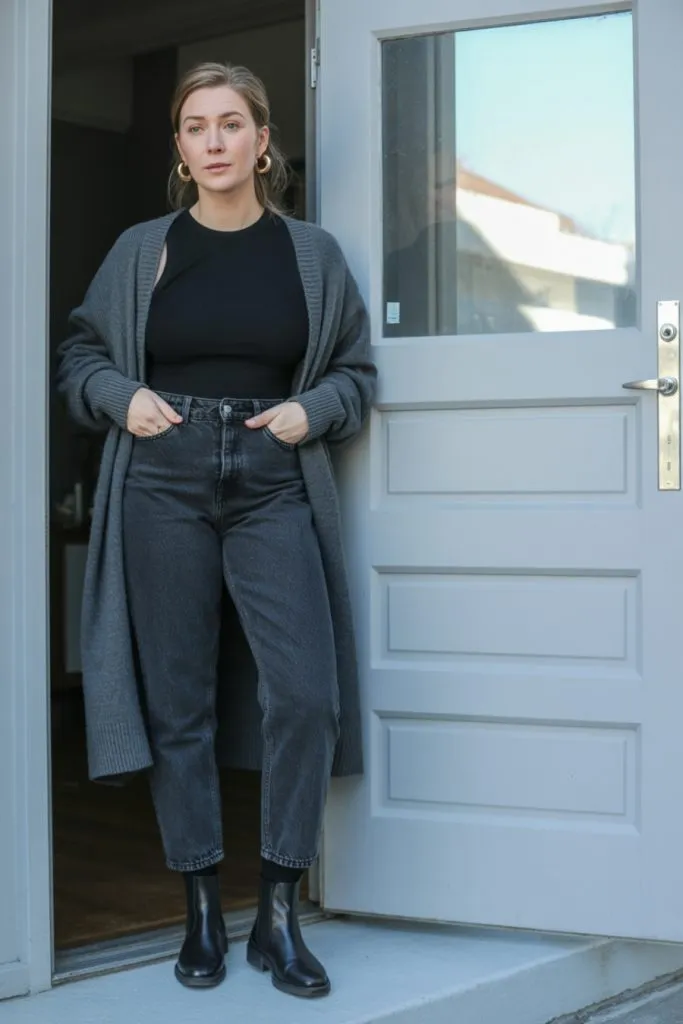 Woman in grey cardigan, black top, and black straight jeans with Chelsea boots standing by a door