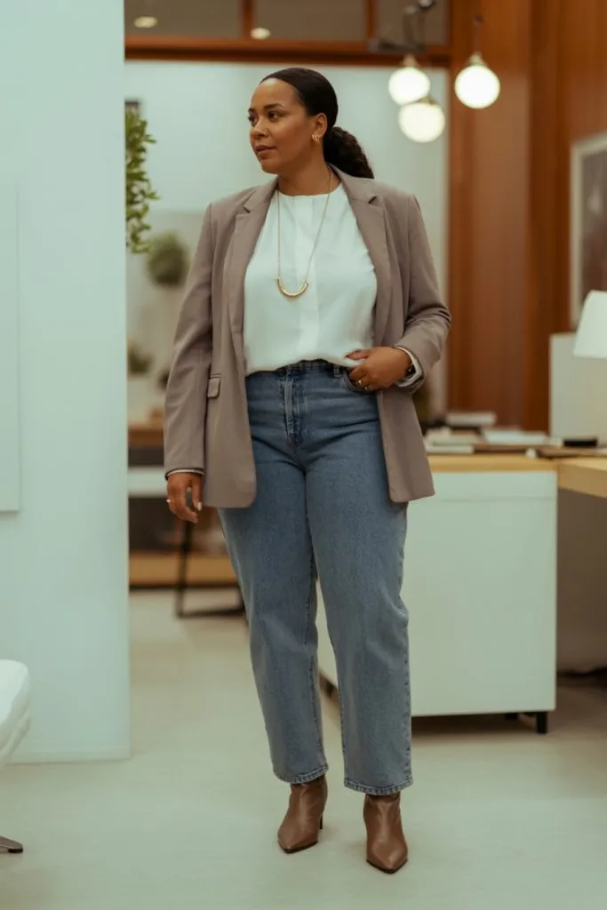Woman in straight jeans, white blouse, taupe blazer and brown heeled boots standing in office setting