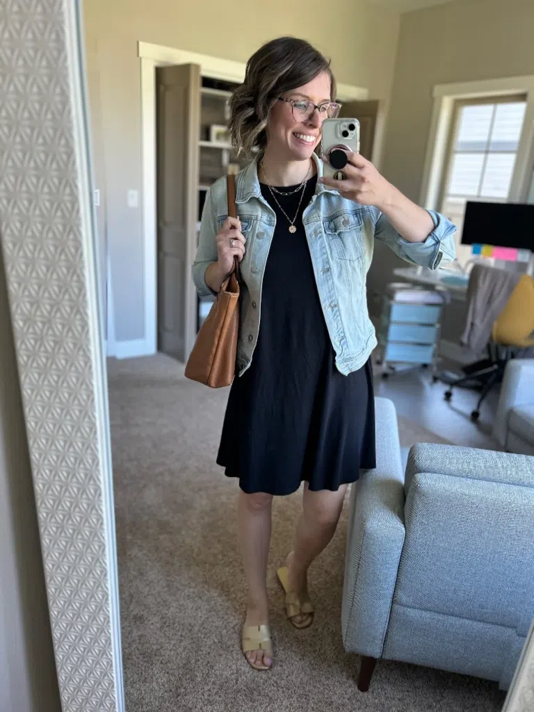 Woman wearing a black dress with a light denim jacket and tan sandals in a mirror selfie