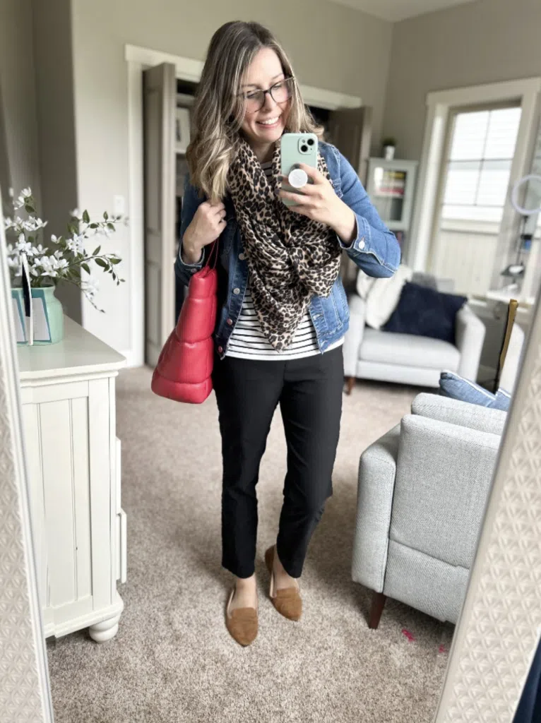 Woman wearing black pants with a striped top, denim jacket, leopard scarf, and loafers in a mirror selfie